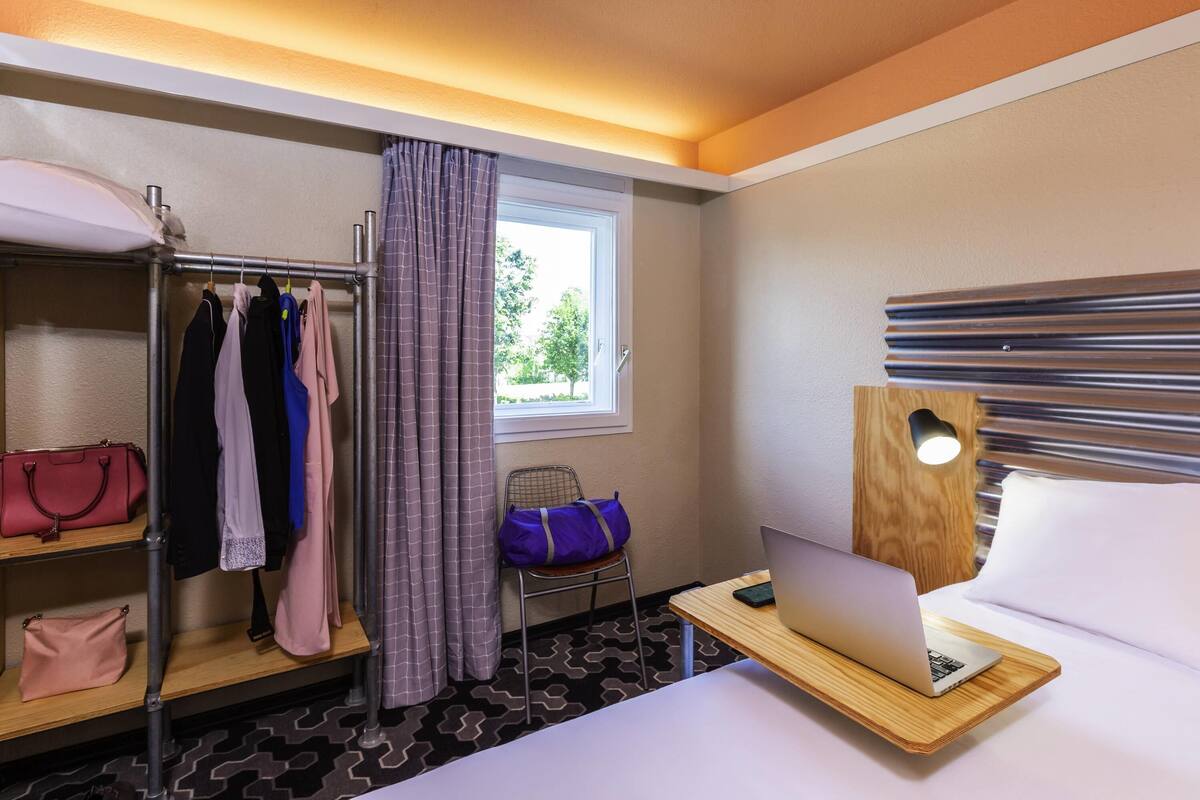 A hotel room with a bunk bed, a desk with a laptop, a window with curtains, and a bed with a pillow.