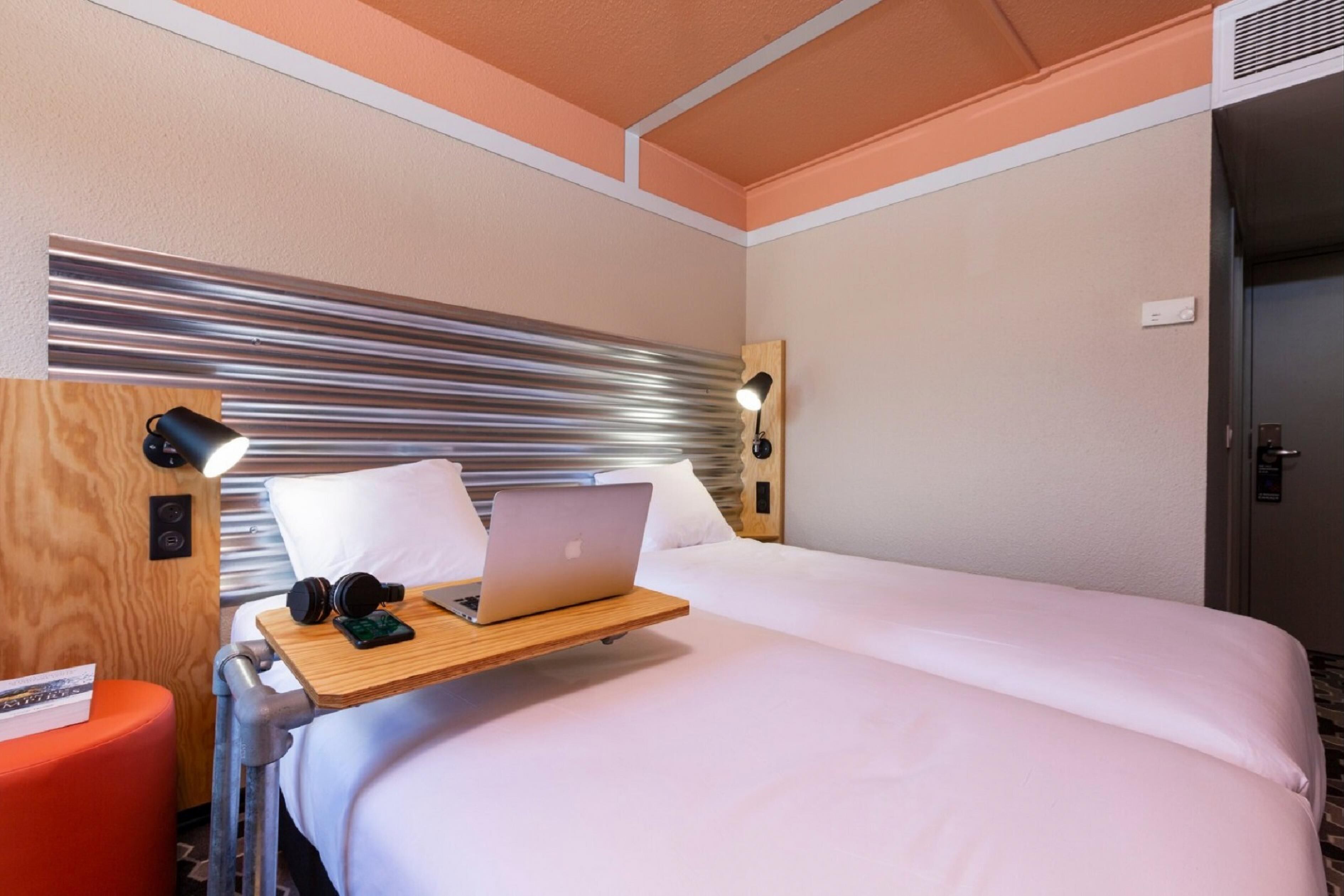 A hotel room with a bed, a desk with a laptop, headphones, and a book.