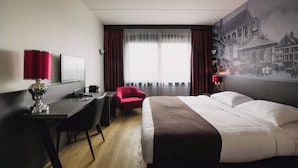 Habitación de hotel con cama, escritorio, silla y un ventanal con cortinas.