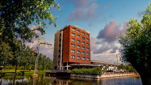 A multi-story hotel with a prominent sign, situated beside a calm body of water, surrounded by greenery.