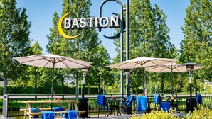 An outdoor seating area with tables and umbrellas, a sign reading "BASTION," and a view of trees and a body of water.
