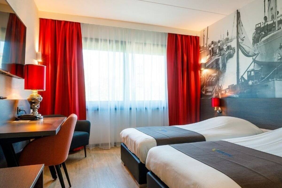 Une chambre d’hôtel avec deux lits, un bureau, une chaise et une grande fenêtre avec des rideaux rouges.