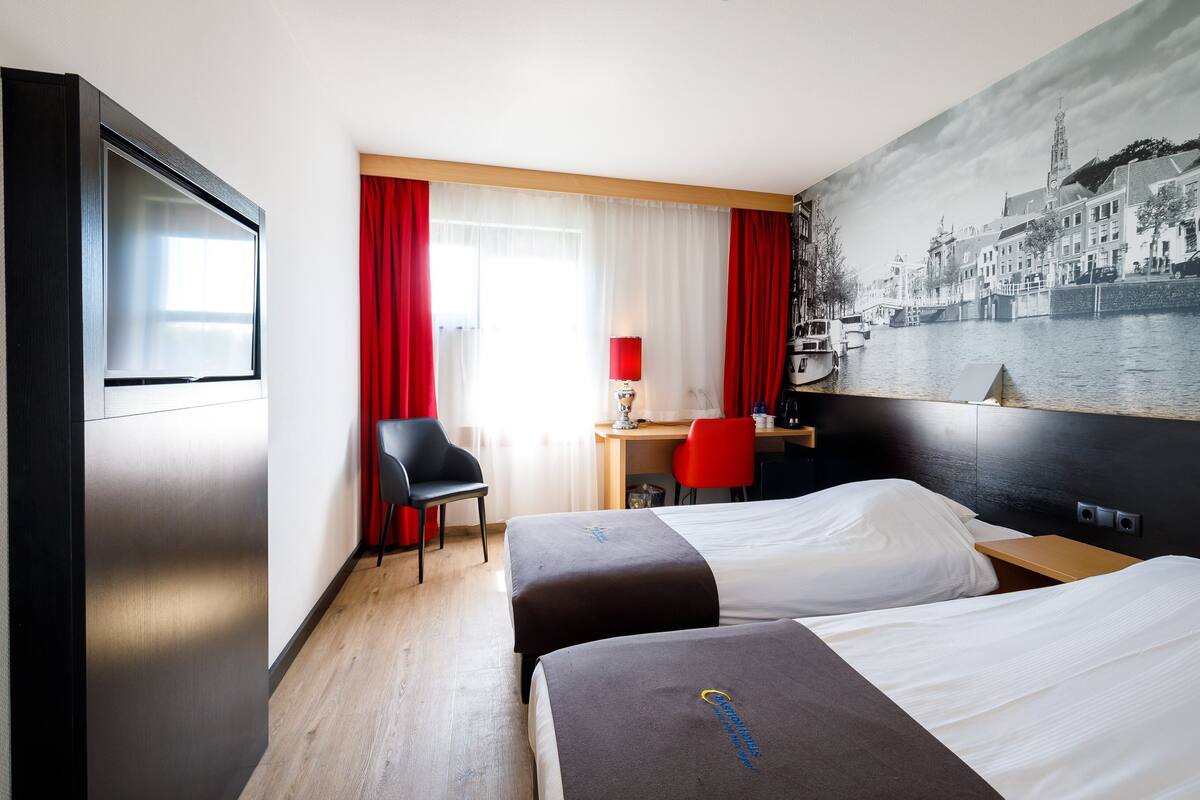Une chambre d’hôtel avec deux lits, un bureau avec une chaise, une fenêtre avec des rideaux rouges et une fresque représentant un paysage urbain sur le mur.