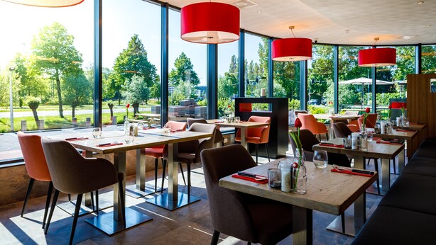 A modern restaurant with large windows, red pendant lights, and a view of greenery outside.