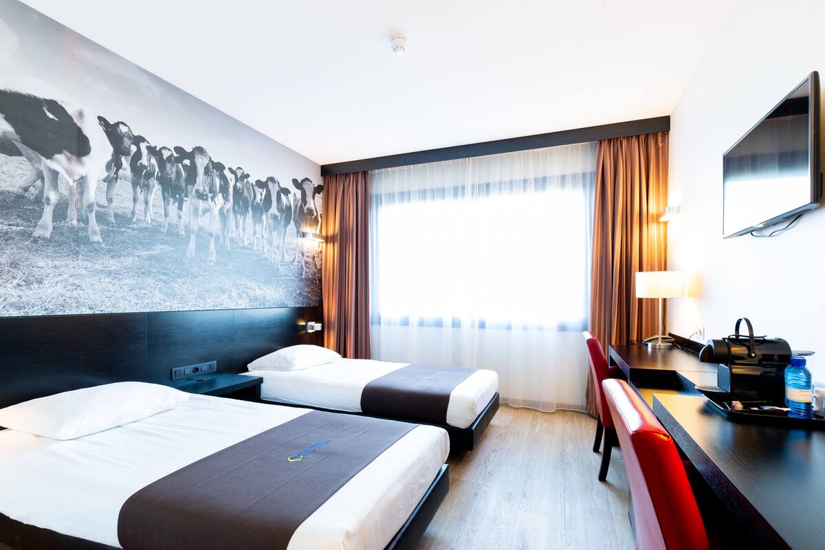 A hotel room with two beds, a desk, a chair, a TV, and a large painting on the wall.