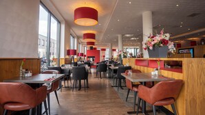 Un restaurante moderno con techos altos, lámparas colgantes rojas y detalles de madera. Las mesas están dispuestas con sillas negras y un ventanal grande ofrece vistas a la calle.