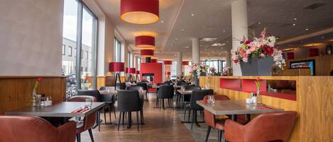 Restaurante moderno com pé-direito alto, luminárias pendentes vermelhas e detalhes em madeira. Mesas dispostas com cadeiras pretas, e uma grande janela oferece vista para a rua.