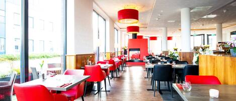 A modern dining area with red and black chairs, wooden tables, and large windows offering a view of the outside.
