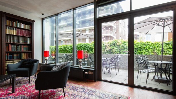 Una habitación con un ventanal grande, una estantería, un sillón negro, una mesa con sillas y una lámpara roja.