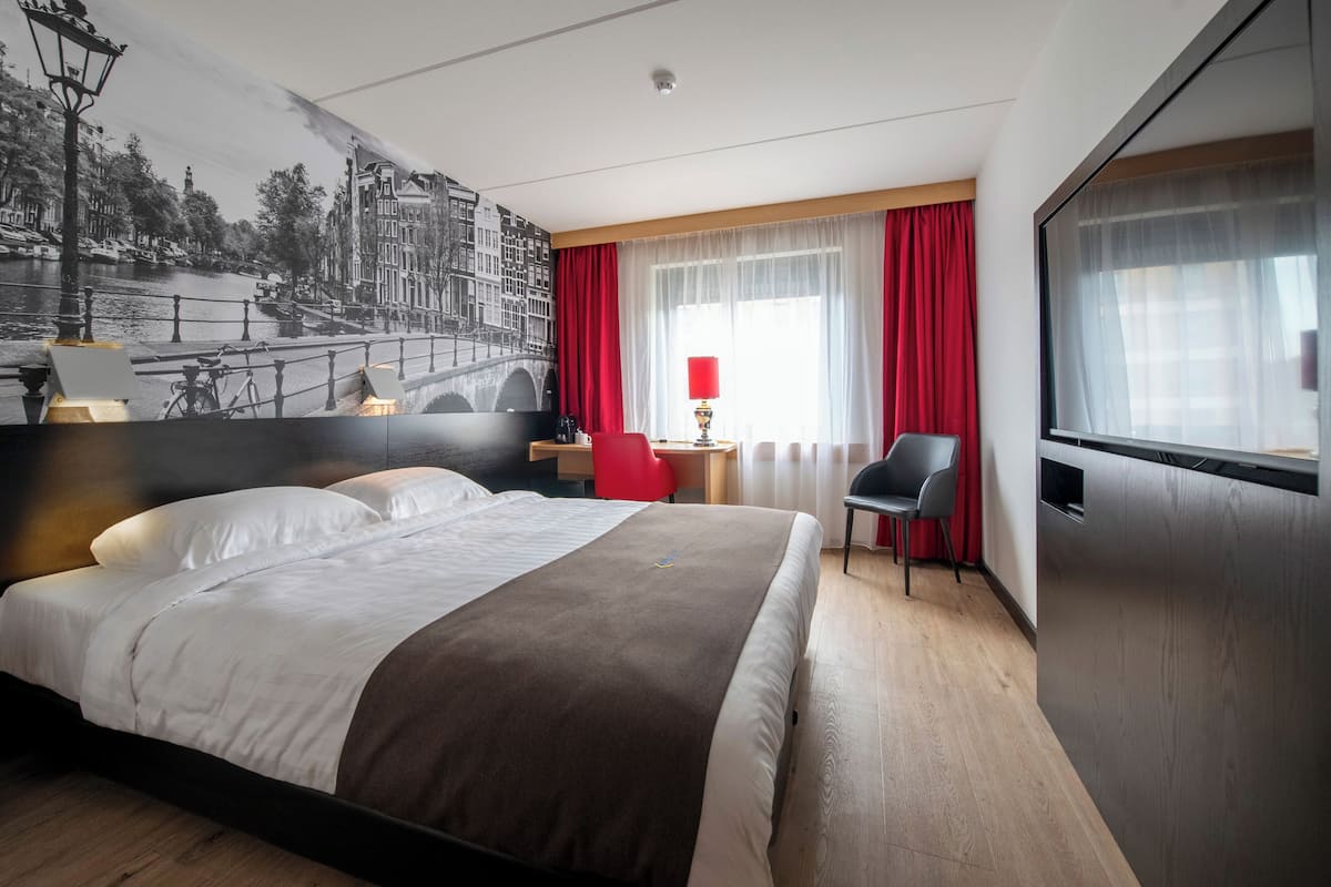 Una habitación de hotel con una cama grande, un escritorio, una silla, un televisor y un mural de un paisaje urbano.