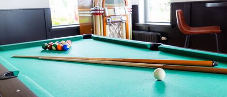 A pool table with balls and cues, a jukebox, and a chair in a room with windows.