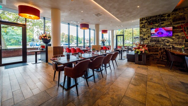 A modern restaurant with large windows, red chairs, and a TV on the wall. - Bastion Hotel Tilburg (Tilburg)