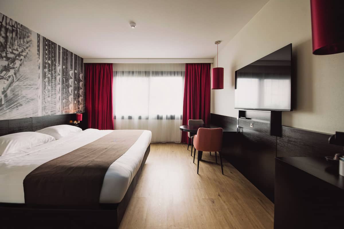 Habitación de hotel con una cama grande, un escritorio, una silla, un televisor y una cortina roja.