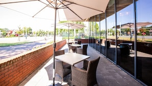 An outdoor patio with wicker furniture and large umbrellas, overlooking a street with a brick wall and greenery. - Bastion Hotel Tilburg (Tilburg)