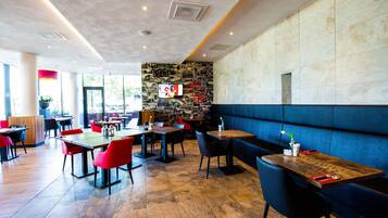 Een modern restaurant met houten tafels en rode stoelen, een stenen muur met een televisie, en een blauwe zithoek.