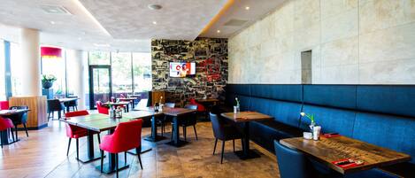 Un restaurante moderno con mesas de madera y sillas rojas, una pared de piedra con un televisor, y una banqueta azul.