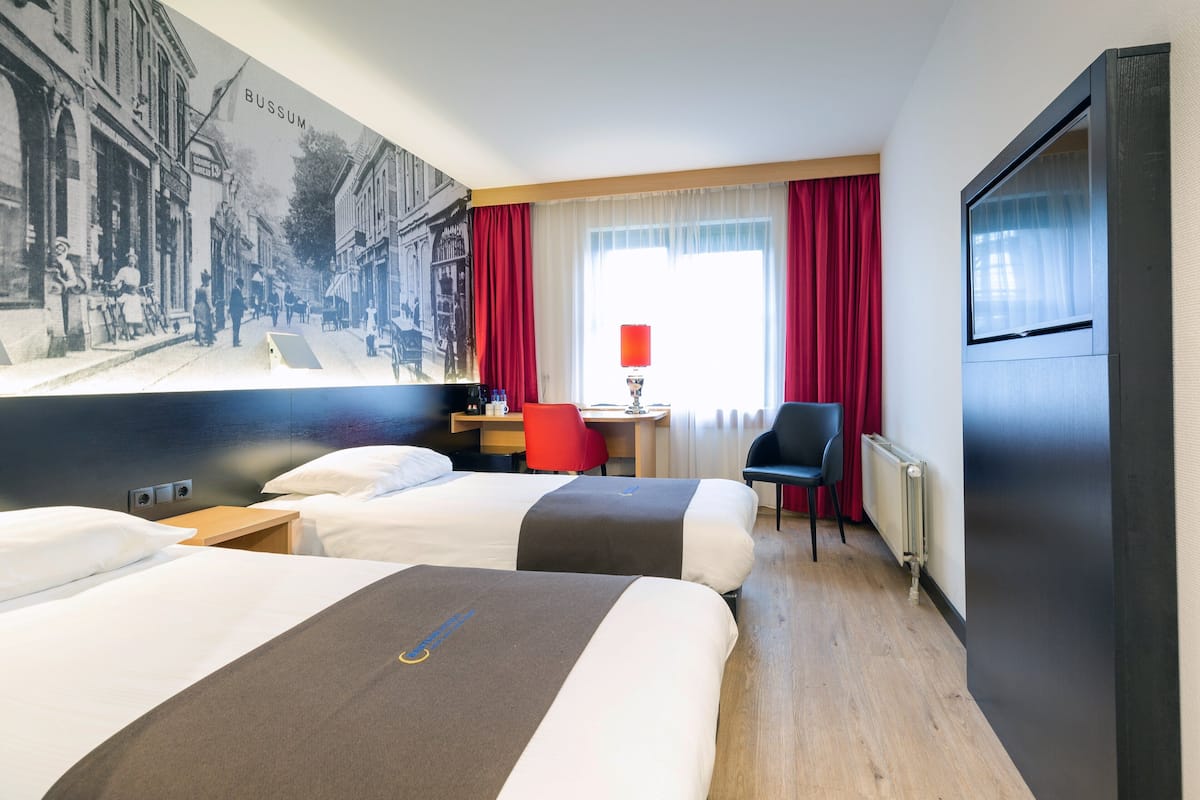 Habitación de hotel con dos camas, un escritorio, una silla, un ventanal grande con cortinas rojas y un televisor instalado en la pared.