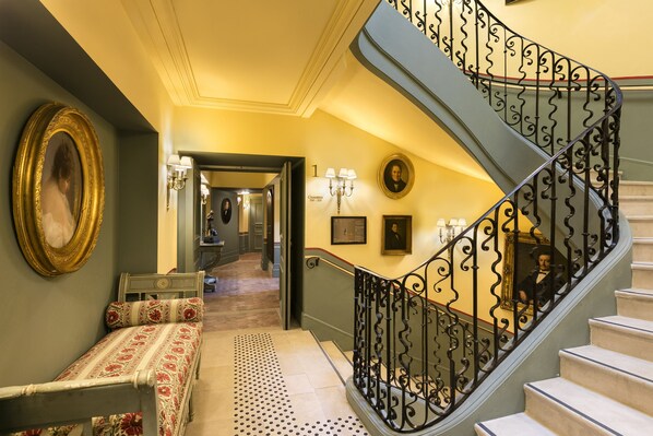 A grand staircase with ornate black railings, a patterned bench, and a hallway leading to a room with a large mirror and wall-mounted lamps. - Airelles, La Bastide de Gordes (Gordes)