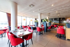 Un restaurante moderno con sillas rojas y mesas blancas, ventanas amplias y plantas decorativas.