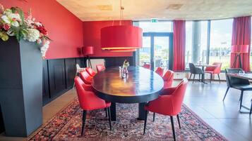 Een moderne vergaderruimte met een ronde tafel, rode stoelen, een rode hanglamp en een groot raam met gordijnen.