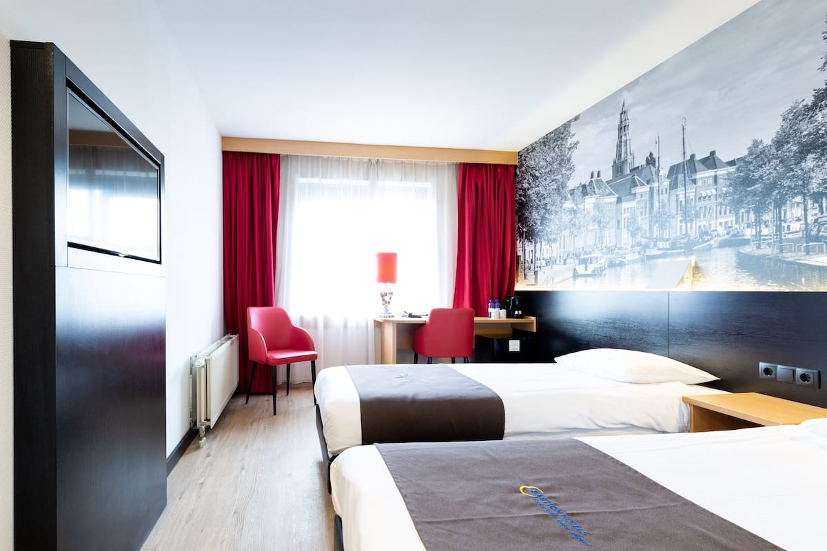 A hotel room with two beds, a desk, a chair, a TV, and a large cityscape painting on the wall.