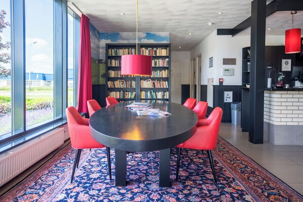A modern meeting room with a round table, red chairs, a large bookshelf, and a view through large windows.