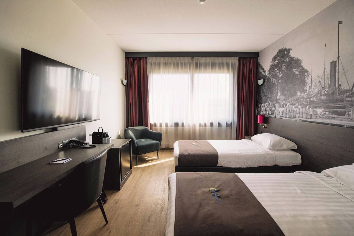 Habitación de hotel con cama, escritorio, silla, televisor y ventana con cortinas.