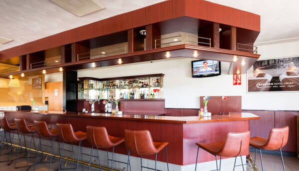 A bar area with a wooden counter, multiple bar stools, a television mounted on the wall, and a visible liquor display.
