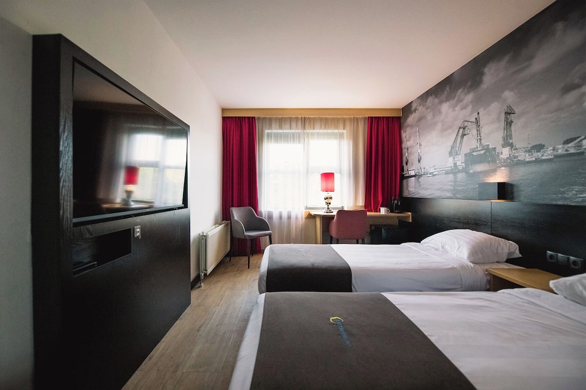 Habitación de hotel con dos camas, un escritorio, una silla, un televisor y un cuadro de un paisaje urbano.