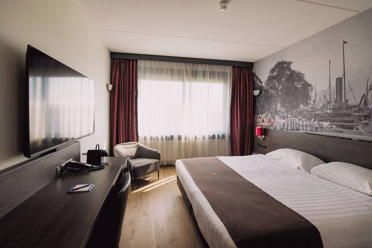 Habitación de hotel con cama, escritorio, silla, televisor y ventana con cortinas.