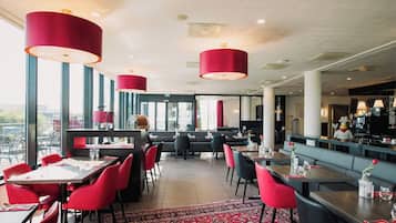 Een restaurant met rode stoelen, tafels en grote rode hanglampen. Er zijn grote ramen en uitzicht op de stad.