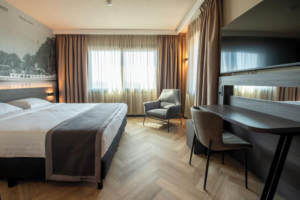 Una habitación de hotel con cama, escritorio, silla y un ventanal con cortinas.