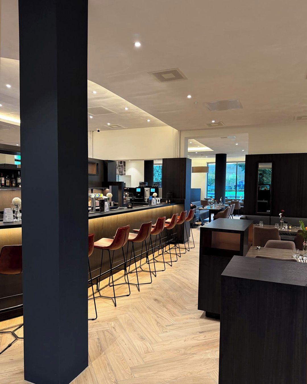Eine moderne Bar mit einer langen Theke und mehreren Hockern, eine Sitzecke mit Tischen und Stühlen sowie eine im Hintergrund sichtbare Küche.