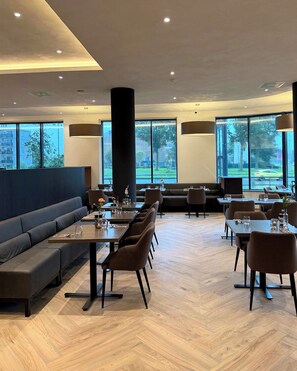 A modern dining area with high tables and chairs, a long bench, and large windows offering a view of the outdoors. - Bastion Hotel Dordrecht Papendrecht (Dordrecht)