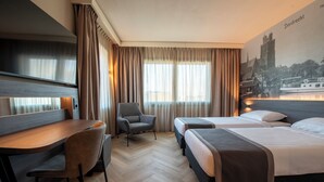 Hotellihuone, jossa on työpöytä, kaksi sänkyä, tuoli ja suuri ikkuna, jossa on verhot.