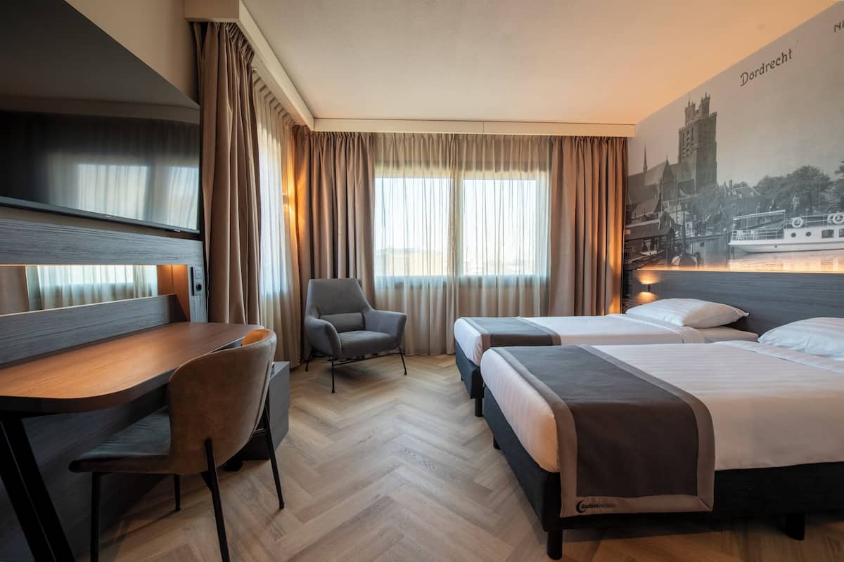 Habitación de hotel con escritorio, dos camas, una silla y un ventanal con cortinas.