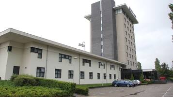 Un bâtiment moderne à la façade blanche, doté d’une haute tour centrale et d’un parking où sont stationnées plusieurs voitures.