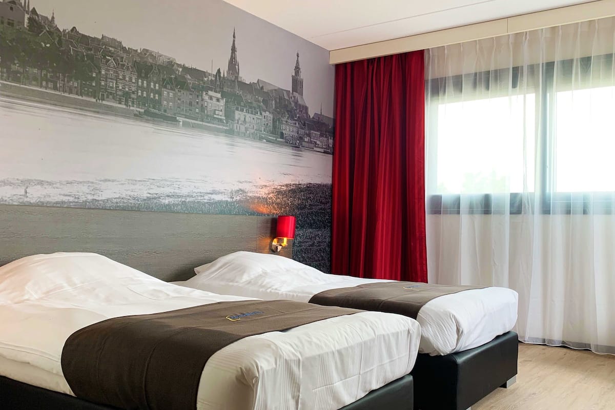 Une chambre d’hôtel avec deux lits, une fresque représentant un paysage urbain sur le mur, un rideau rouge et une fenêtre avec des rideaux en voile.