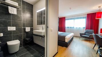 A modern hotel room with a bathroom, a bed, a desk, and a chair.