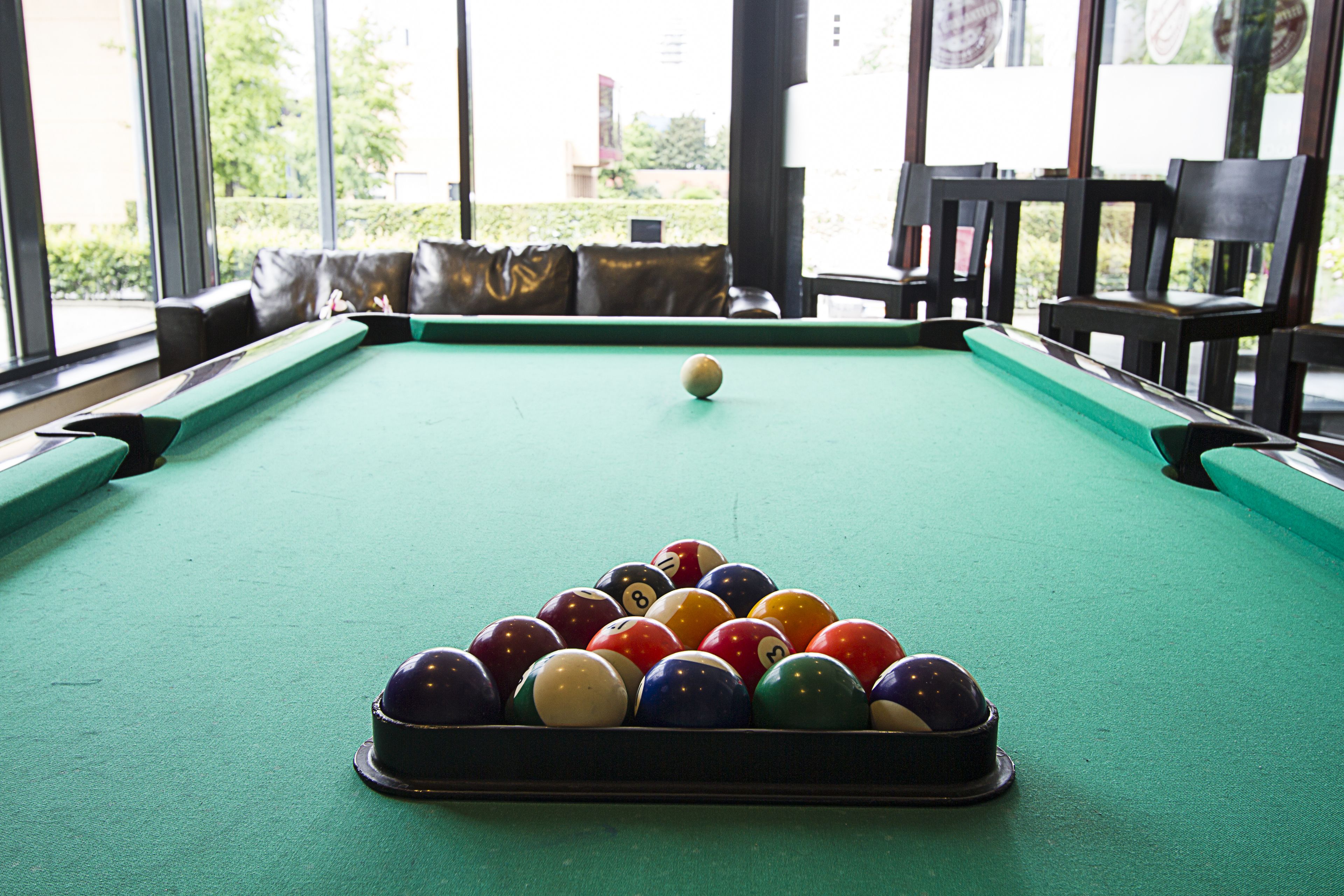 Une table de billard recouverte d’un drap vert, entourée de chaises noires, dans une pièce dotée de grandes fenêtres.
