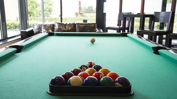 Une table de billard recouverte d’un drap vert, entourée de chaises noires, dans une pièce dotée de grandes fenêtres.