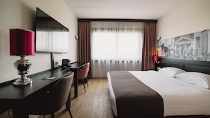 Una habitación de hotel con cama, escritorio, silla y un ventanal con cortinas.