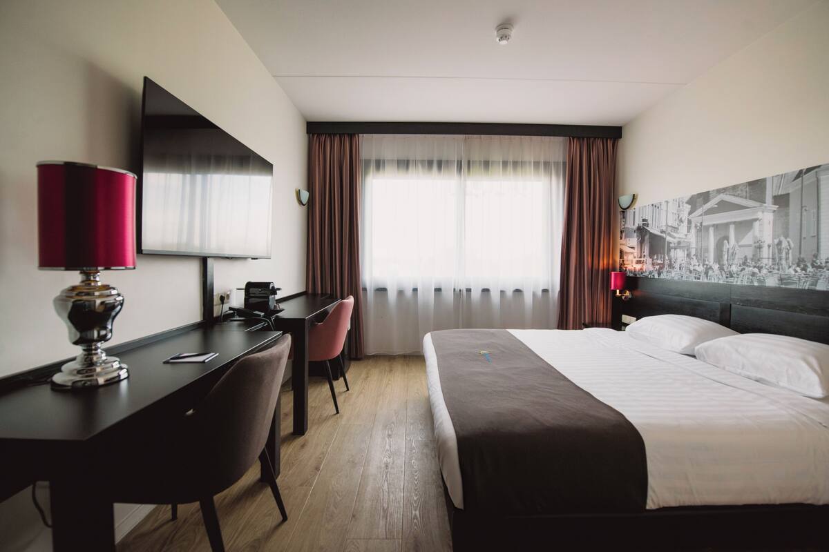 A hotel room with a bed, desk, chair, and a large window with curtains.