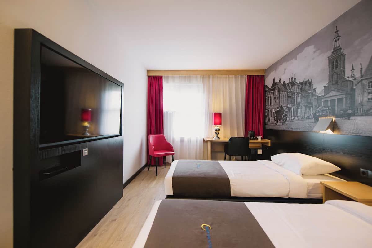 A hotel room with a large flat-screen TV, two beds, a desk with a chair, and a mural of a cityscape on the wall.