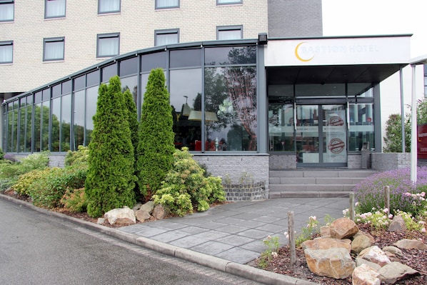 A modern building with a glass facade, a sign reading "Bastion Hotel," and a landscaped entrance area with shrubs and rocks. - Bastion Hotel Zoetermeer (Zoetermeer)
