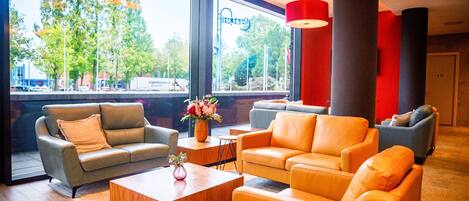 A modern lobby with leather sofas, a wooden coffee table, and large windows offering a view of trees and buildings.