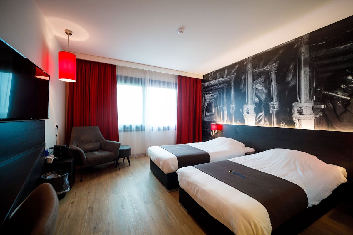 A hotel room with two beds, a desk, a chair, a red pendant light, and a large abstract painting on the wall.