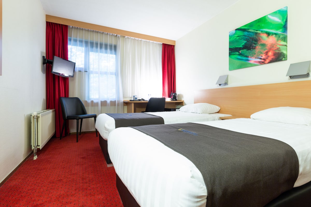 A hotel room with two beds, a desk, a chair, a TV, and a painting on the wall.