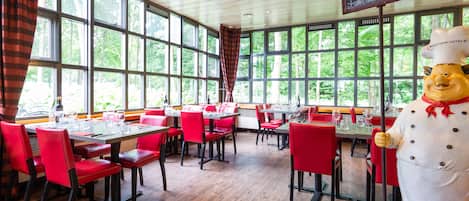 A restaurant with red chairs and tables, large windows, and a chef statue.