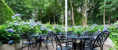 An outdoor patio with black metal tables and chairs, surrounded by lush greenery and hydrangea flowers.
